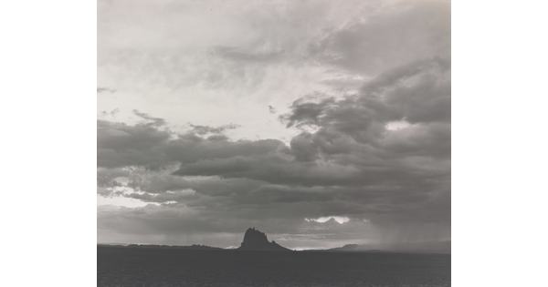 A black-and-white photograph of a dramatic light and dark sky with some striated clouds and a rock formation in the distance.