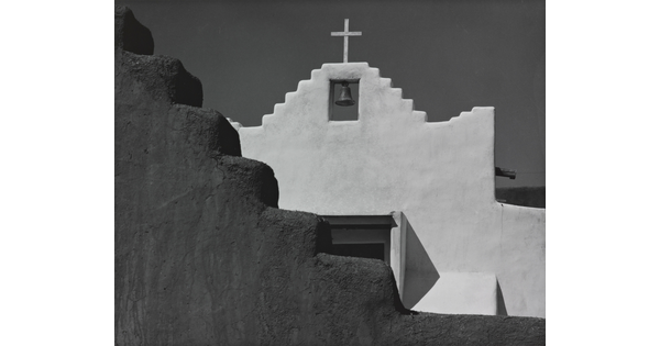 A black-and-white photograph of a terraced adobe bell tower with a cross on top, partially hidden by a terraced adobe wall.