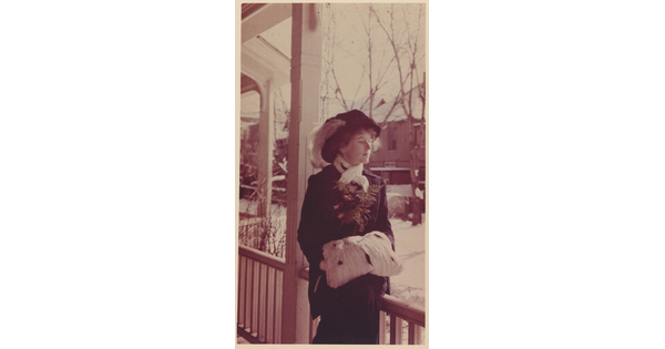 A sepia-toned portrait photograph of a White woman wearing a hat with a feather in it, a scarf, coat, hands tucked into a muff, standing on a porch.