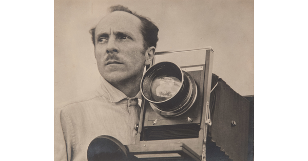A sepia-toned portrait photograph of a balding White man with a mustache beside a large-format camera.