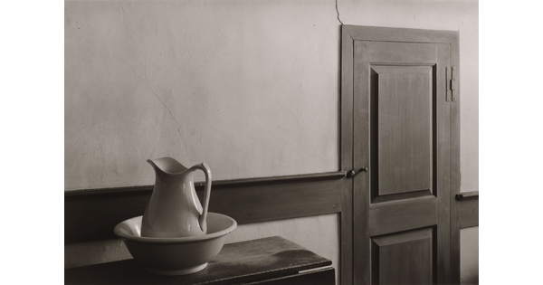 A black-and-white photograph of a ceramic pitcher and wash basin on a table next to a wooden door.
