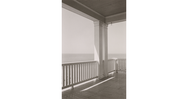A black-and-white photograph of porch with a white-slatted railing overlooking the ocean.