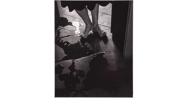 A black-and-white photograph of a person's bare feet, toes stretched up off the tile floor to avoid puddles.