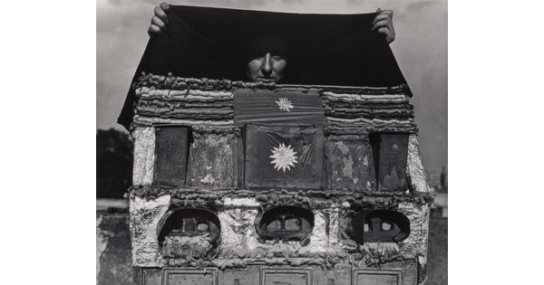 A black-and-white photograph of a person holding a black cloth above what looks like a homemade dollhouse, but instead of windows, it has stereoscopes embedded in it.