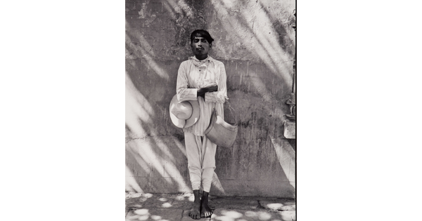 A black-and-white photograph of a dark-skinned man in light-colored shirt and pants, holding a hat under one crossed arm and a bag on the other, leaning against a wall.