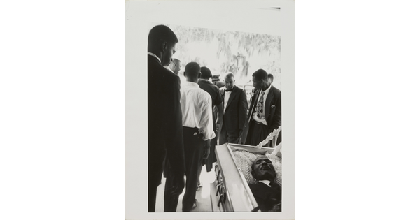 A black-and-white photograph of Black men in suits filing by an open casket.