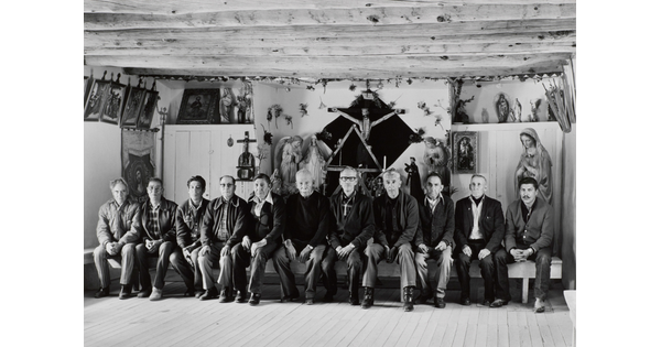 A black-and-white photograph of a group of men, most of them older, seated on a wood bench in front of many religious sculptures and paintings.