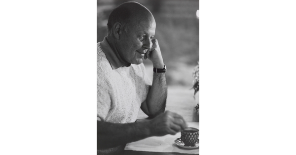 A black-and-white portrait photograph of a bald White man in profile resting his chin on a closed fist as his other hand stirs liquid in a cup.