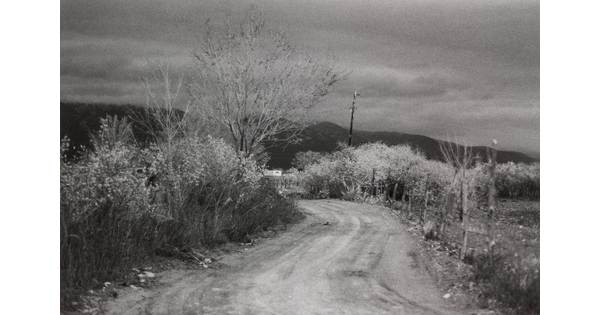 A black-and-white photograph of a curvy dirt road with scrubby vegetation on both sides, distant hills, all under a dark sky.