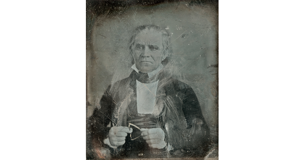 A worn and scratched black-and-white photograph of a seated, older White man in a suit.