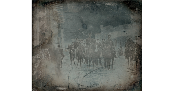 A dark, black-and-white photograph of a group of figures in military uniform on horseback on a street.