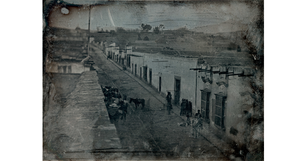 A black-and-white photograph, slightly obscured by surface marks, of a street lined with buildings, people and horses.
