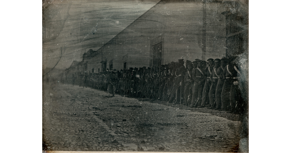 A dark, black-and-white photograph of a group of figures in military uniforms lined up against a building on a cobblestone street.