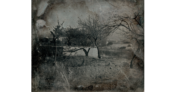 A scratched black-and-white photograph of a wooden cross marking a burial site, surrounded by leafless trees on a dirt hill.