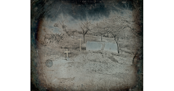 A faded black-and-white photograph of two wooden crosses marking burial sites, surrounded by leafless trees on a dirt hill.