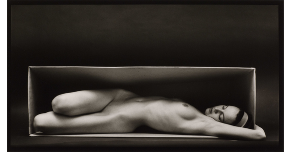 A black-and-white photograph of a nude, fair-skinned woman lying on her back, twisting toward the viewer, in a box.