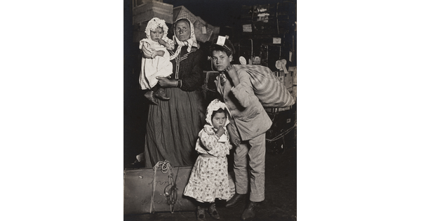 A black-and-white photograph of a light-skinned woman with three children of different ages, standing with suitcases and bags.
