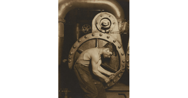 A black-and-white photograph of a muscular man crouched next to a large piece of machinery that he is adjusting with a large wrench.