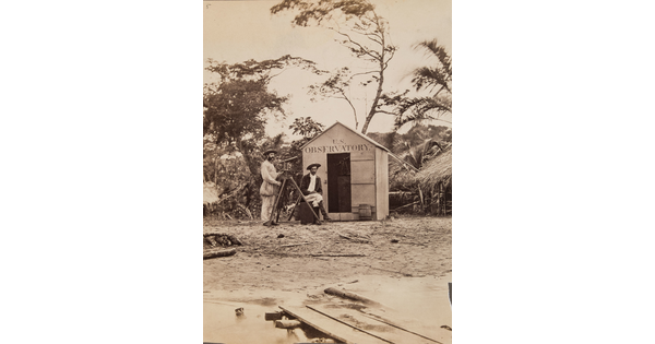 A black-and-white photograph of two men with a telescope standing in front of a small shack with "U.S. Observatory" above the doorway.