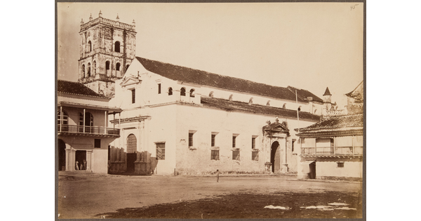 A sepia-toned photograph of dirt forecourt leading to a light-colored two-story structure and an ornate square tower in the background.