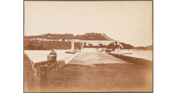 A sepia-toned photograph of a wide stone bridge leading across a river to arched structures on a rocky hillside.