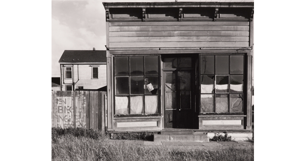 A black-and-white photograph of a wood storefront with cloth covering the windows and doors on the inside; overgrown grass lines the front walk.