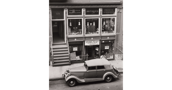 A black-and-white photograph of a vintage car parked on the street outside of a storefront advertising antiques.