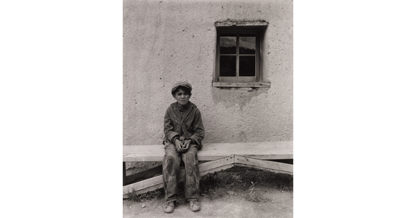 A black-and-white photograph of a squinting medium-skinned boy wearing a cap, jacket, and pants with patches on the knees, seated on a wooden bench in front of an adobe building.