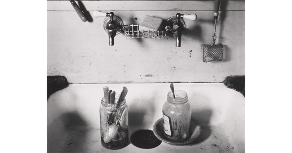A black-and-white photograph of a two-tap sink with two dirty mason jars full of spoons.