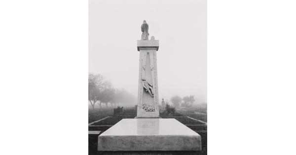 A black-and-white photograph of a tall, vertical stone monument rising behind a flat stone slab in a cemetery.