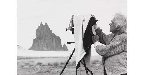 A black-and-white photograph of an older White woman with a box camera on a tripod and a tall rock formation in the background.