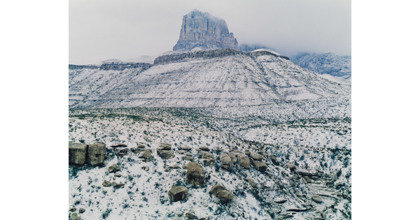 A color photograph of a snowy landscape featuring a tall butte and distant mountains obscured by snowy haze.