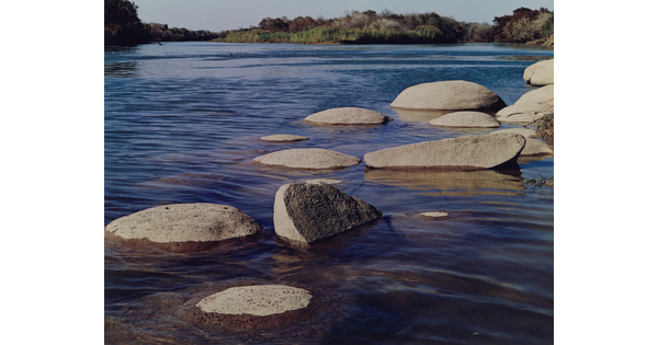 A color photograph of many, large smooth boulders in a river reflecting a blue sky.