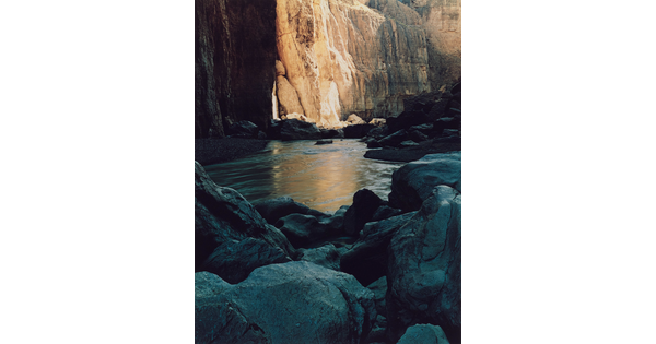 A color photograph of large boulders resting in still water at the base of a cliff.