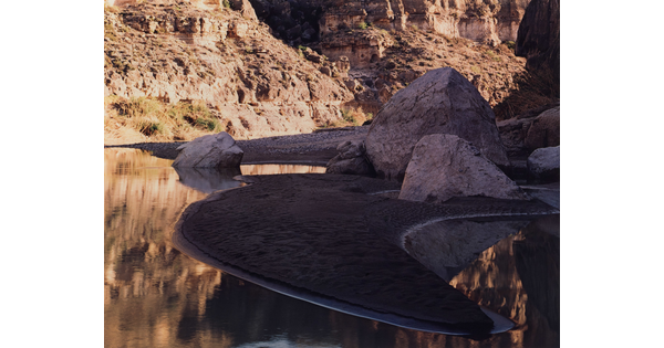 A color photograph of large boulders on a sandy riverside beach and a rocky cliff in the background.