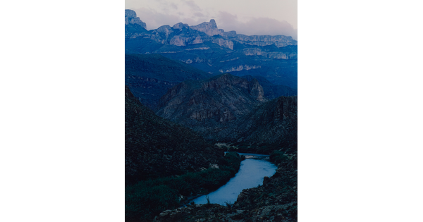 A color photograph of a river in a valley surrounded by blue mountains and cliffs.