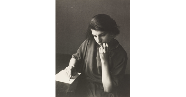 A black-and-white photograph of a young White woman seated at a table writing on a piece of paper.