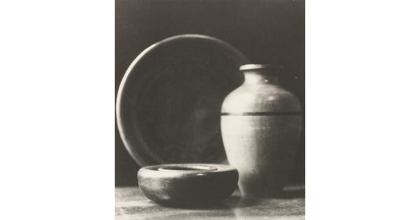 A black-and-white photograph of a short wood bowl, a large pear-shaped vase, and a standing plate behind them.