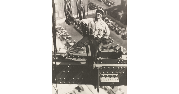 A black-and-white photograph of a smiling White man standing on a beam holding onto a chain, wearing a newsboy cap, cars in parking lots far below.