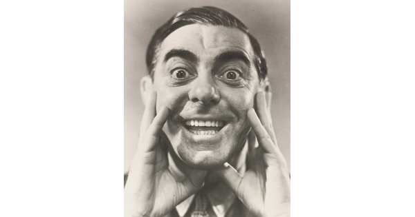 A black-and-white portrait photograph of a White man, hair slicked back, hands framing his cheeks, mouth open in a wide smile.