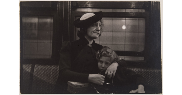 A black-and-white photograph of a White woman with her arms around a child; both seated on an upholstered seat with windows behind them.