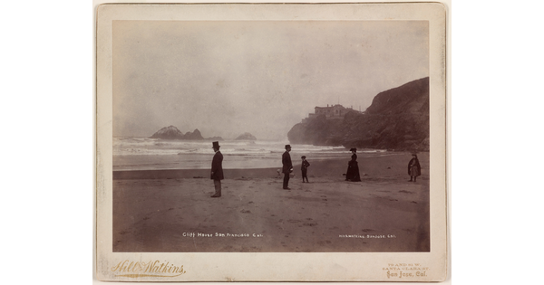 A black-and-white photographs of formally-dressed people scattered along a flat beach and calm shoreline; in the background, a building sits atop a high cliff.