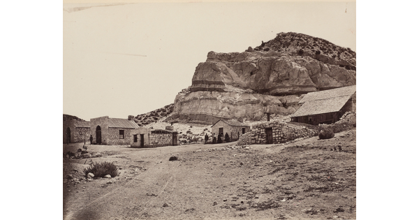 A black-and-white photograph of a group of small stone buildings at the base of a cliff in a desert landscape.