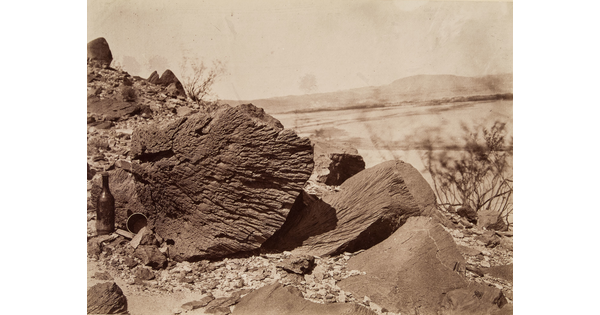 A black-and-white photograph of a striated rock and a glass bottle standing next to it in a desert landscape.