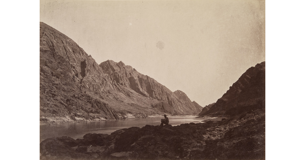A black-and-white photograph of a person sitting on a rock next to a calm river surrounded by steep cliffs and mountains.