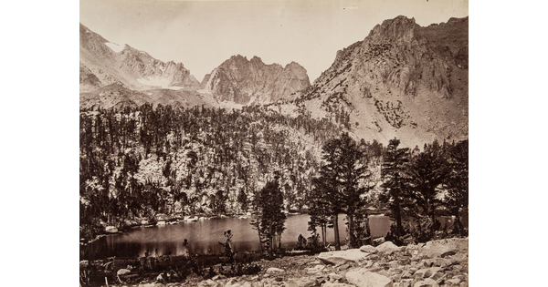 A black-and-white photograph of a lake in a mountain valley surrounded by pine trees and mountains dusted in snow.