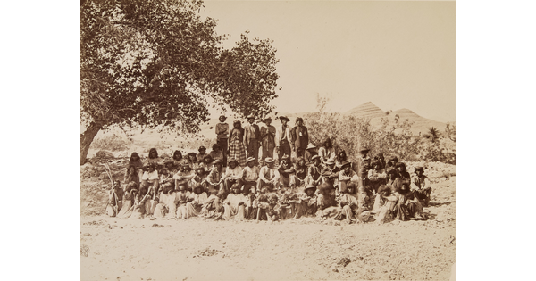 A black-and-white photograph of a large group of Indigenous people, some seated and some standing, under a tree with mountains in the distance.