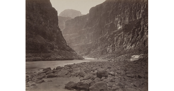 A black-and-white photograph of a calm river at the rocky bottom of a canyon.