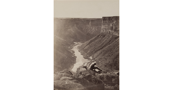 A black-and-white photograph of a person sitting on rocks above a river running through a canyon surrounded by tall cliffs.