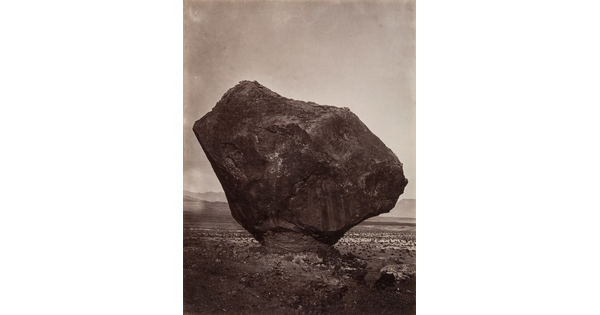 A black-and-white photograph of a large rock formation balanced on a narrow pedestal in a desert.